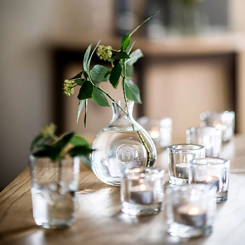 The Anna Viktoria Glass Dala Horse Vase, mouth-blown in Portugal, holds vibrant green leaves alongside small candle holders on a wooden table.