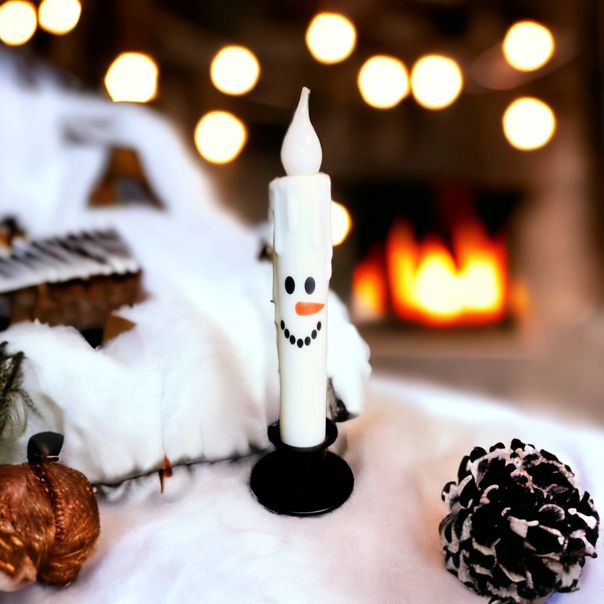 The Candle: Snowman Face Christmas Battery Operated Timer Taper Candles feature a snowman-shaped design with a black base, surrounded by snowy decorations, a pine cone, and an acorn. Blurred warm lights and the glow of a cozy fireplace fill the background.