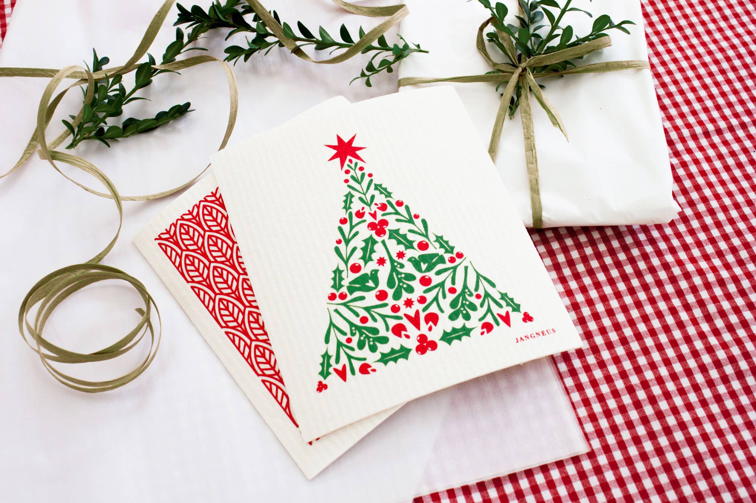 Christmas cards featuring a festive tree design are set on a red and white checkered tablecloth alongside Scandinavian-style Dish Cloths named "Dish Cloth - Christmas Tree - Red." A green ribbon and decorative vine complete the cozy atmosphere, blending practicality with holiday charm.