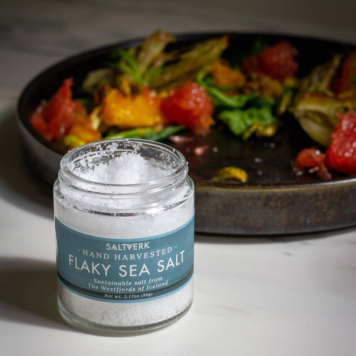 A SALTVERK Gift Set—featuring Flaky Sea Salt, Lava Salt, Birch Smoked Salt, and Arctic Thyme Salt—rests on a marble surface, with a blurred plate of salad and grapefruit segments in the background.