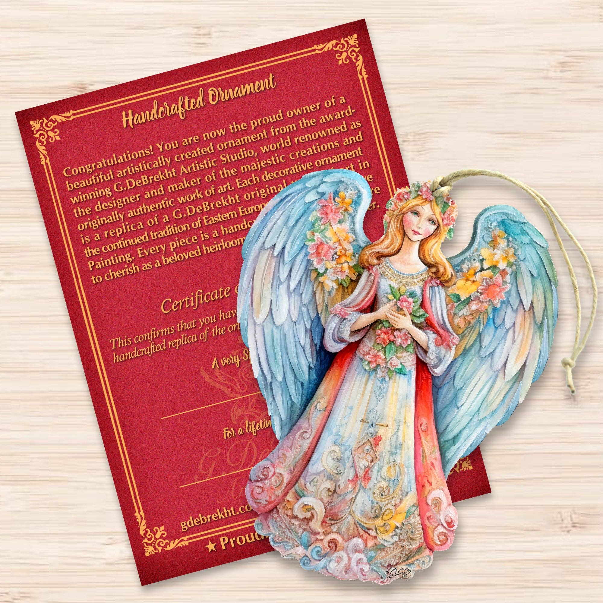 A vibrant "Ornament: Angel Blue Floral" by Debrekht Nativity Holiday, adorned with floral details, is displayed on a red certificate of authenticity, highlighting its distinctiveness as a handcrafted keepsake.