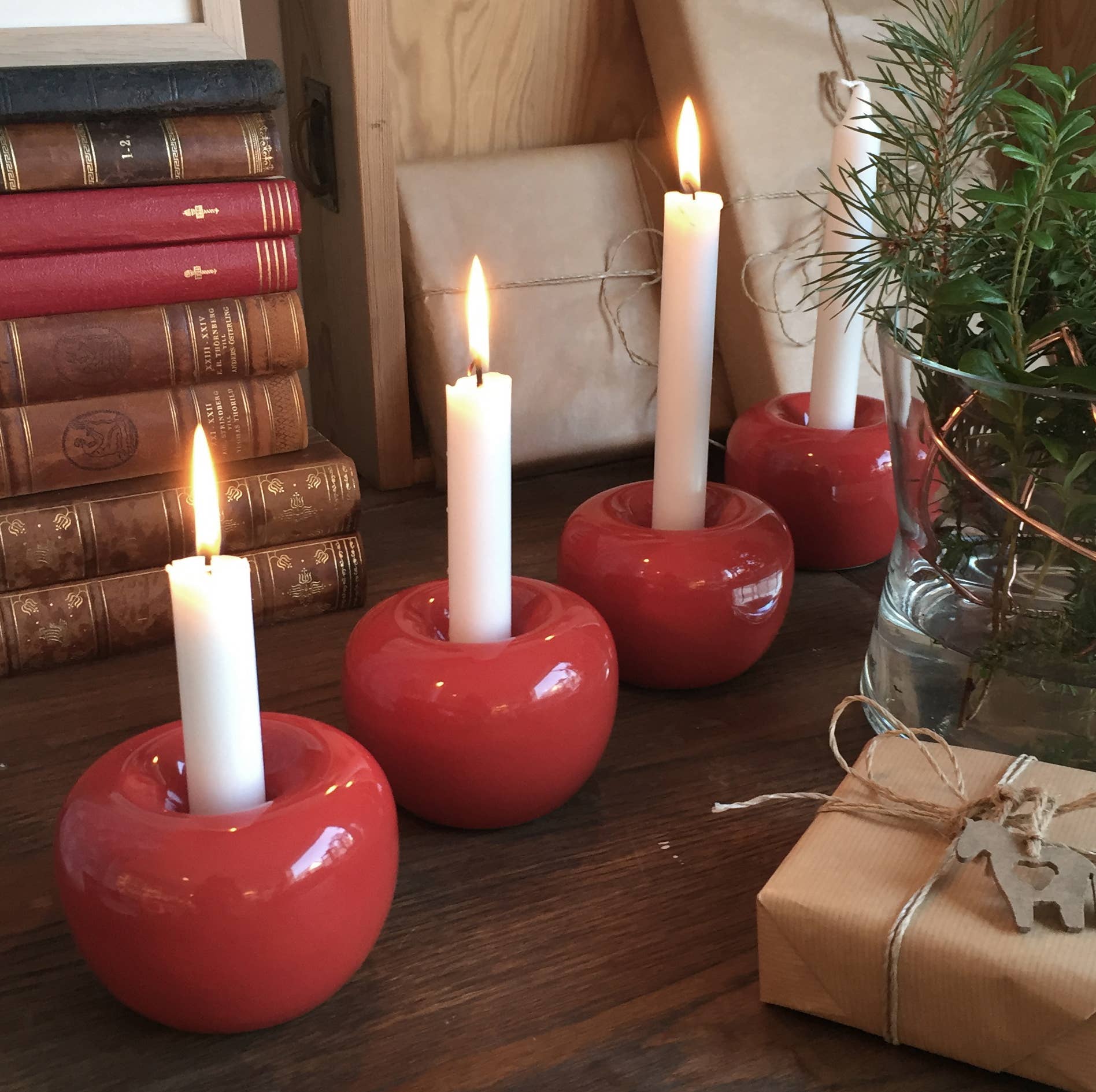 Four lit white candles in Swedish Shiny Red Apple Taper Candle Holders on a wooden table, surrounded by stacked books, wrapped gifts, and a vase with green branches.