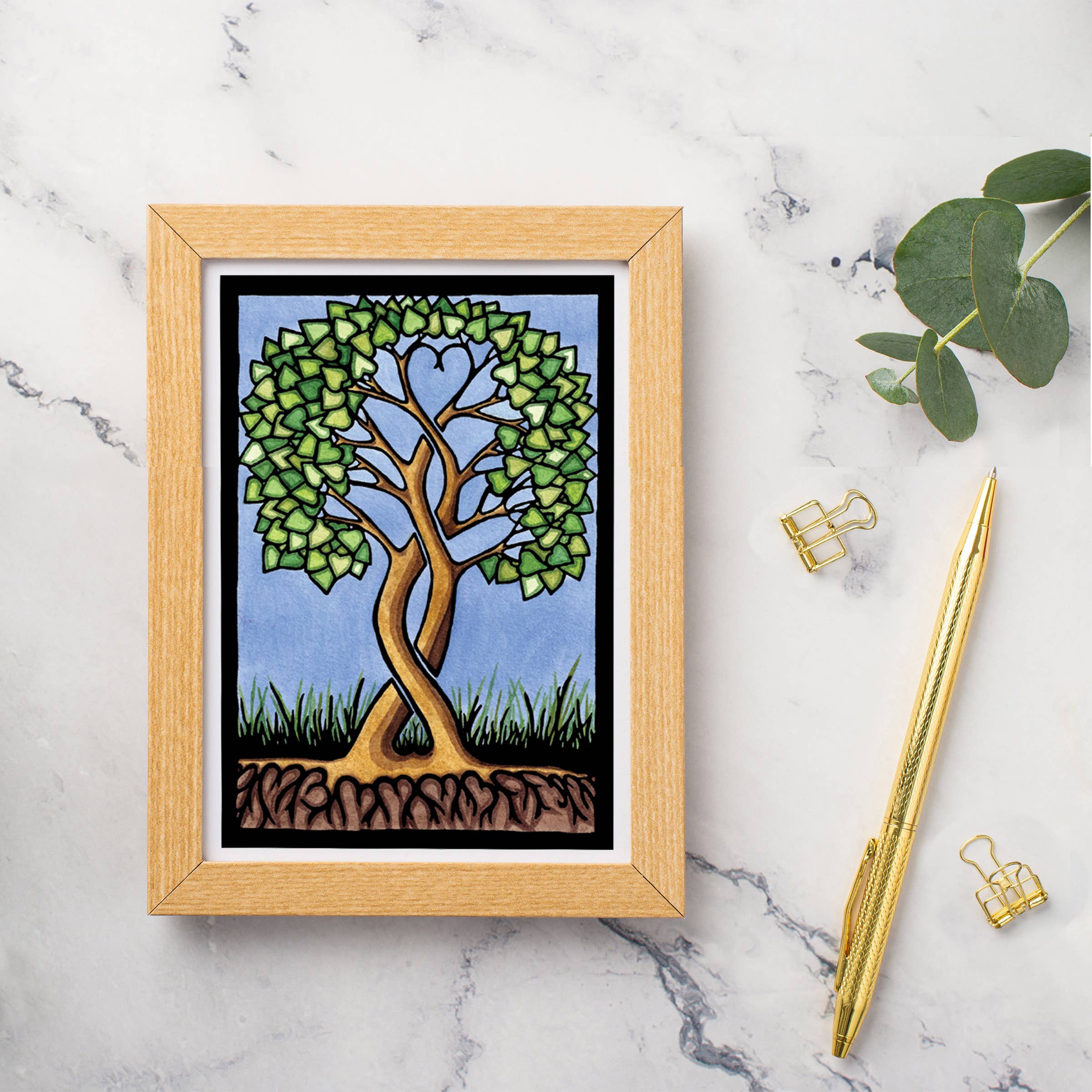 Card: Together Forever Tree by Sarah Angst (5x7) features a stylized tree with heart-shaped foliage. Displayed on marble with a gold pen, paper clips, and eucalyptus leaves. Made in America.