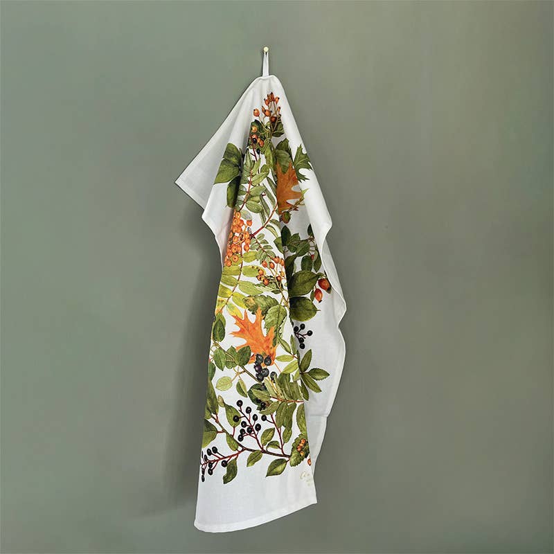 The Tea Towel: Autumn - 100% organic cotton tea towel by Kirsten Tind Koustrup & Co hangs against a plain green wall, its white fabric adorned with a green and orange leaf pattern that adds charm to the kitchen décor. Crafted from organic cotton, it embodies both style and sustainability.