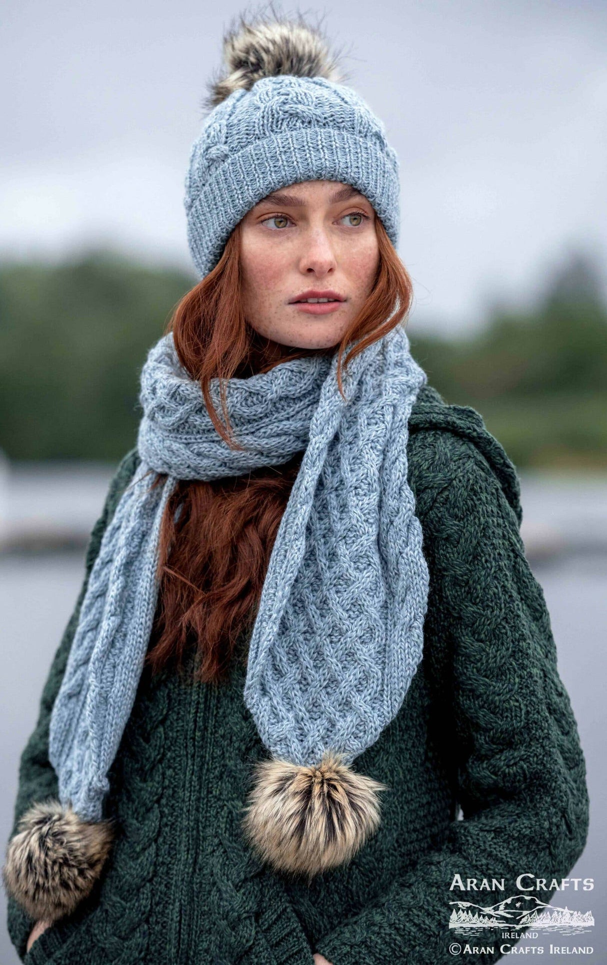A woman with long red hair wears the Chunky Cable Hat - Mermaid (One Size), made in Ireland with faux fur pompoms, along with a scarf and a green merino wool cable-knit sweater, standing outdoors.