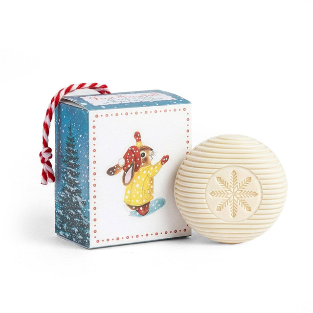 A round soap embossed with a snowflake design next to a holiday ornament box featuring a Richard Scarry rabbit in winter clothing.