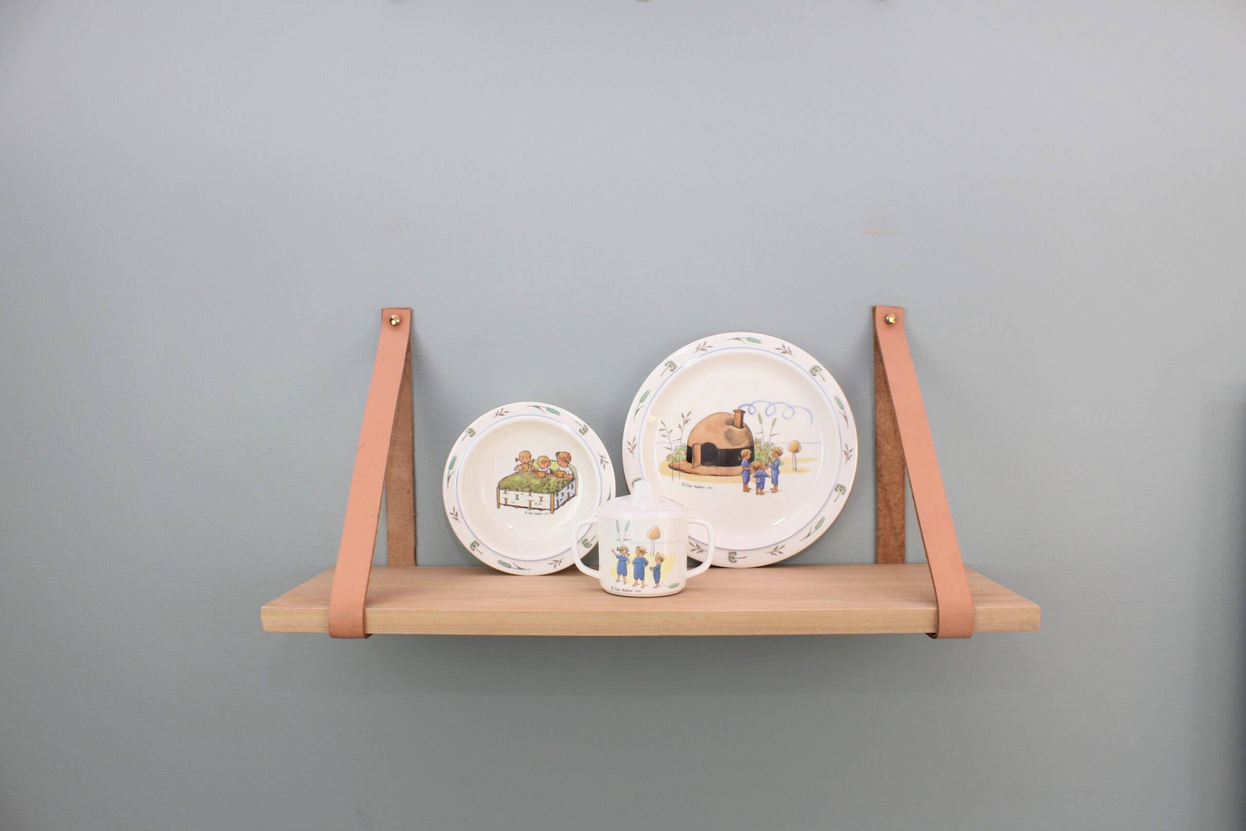 A wooden shelf with leather straps displays three decorative plates and a cup, each showcasing charming illustrations inspired by the Elsa Beskow "Children of Hat Cottage" Dish Set, set against a light gray wall.