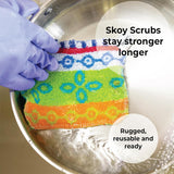A hand in a purple glove holds Skoy Scrub Cloths Good Clean Fun! by Skoy Enterprises in a metal bowl of water. Text: Strong, reusable, eco-friendly—Skoy scrubbers last longer for good clean fun every time.