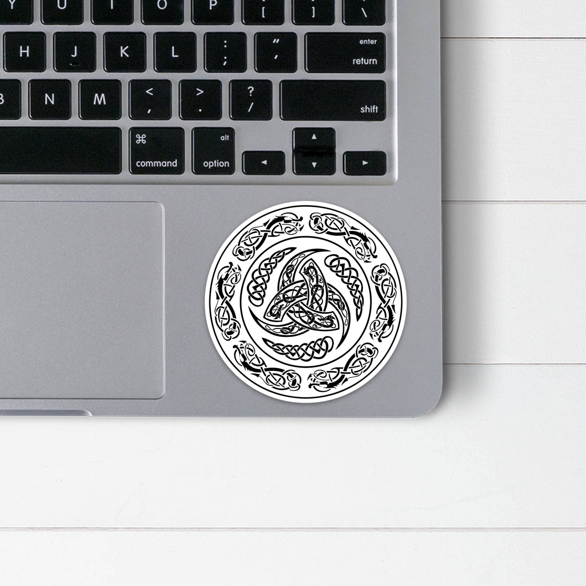 A laptop partially in view, displaying a circular sticker crafted from the highest quality vinyl. This sticker, known as the Viking Shield Sticker, features a black Celtic knot design surrounded by an intricate border near the touchpad.