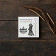A white box labeled Card Pack: Viking Finds by Koustrup & Co is adorned with Viking motifs like a ship, a woman, and a sword. It rests on a wooden surface, surrounded by dried grass.