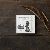 A white box labeled Card Pack: Viking Finds by Koustrup & Co is adorned with Viking motifs like a ship, a woman, and a sword. It rests on a wooden surface, surrounded by dried grass.