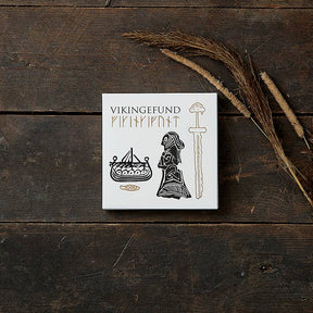 A white box labeled Card Pack: Viking Finds by Koustrup & Co is adorned with Viking motifs like a ship, a woman, and a sword. It rests on a wooden surface, surrounded by dried grass.