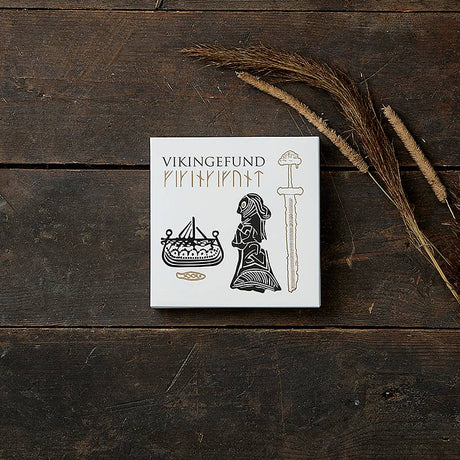 A white box labeled Card Pack: Viking Finds by Koustrup & Co is adorned with Viking motifs like a ship, a woman, and a sword. It rests on a wooden surface, surrounded by dried grass.