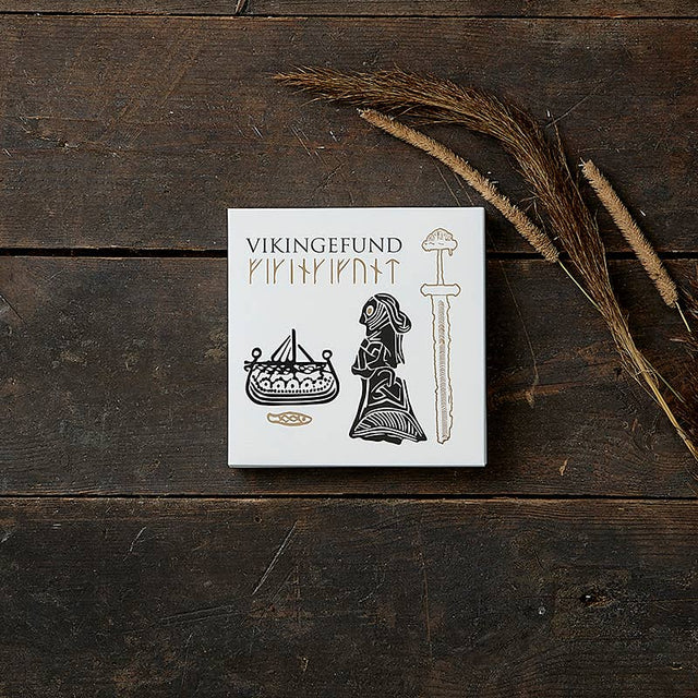 A white box labeled Card Pack: Viking Finds by Koustrup & Co is adorned with Viking motifs like a ship, a woman, and a sword. It rests on a wooden surface, surrounded by dried grass.