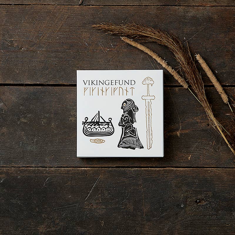 A square box labeled "Card Pack: Viking Square Cardfolder" features intricate Viking motifs, including drawings of a ship, person, and sword on a dark wooden surface. It is beautifully complemented by dried grass stalks and highlighted with bronze foil accents for an authentic touch to this exquisite collection of 8 cards with envelopes.