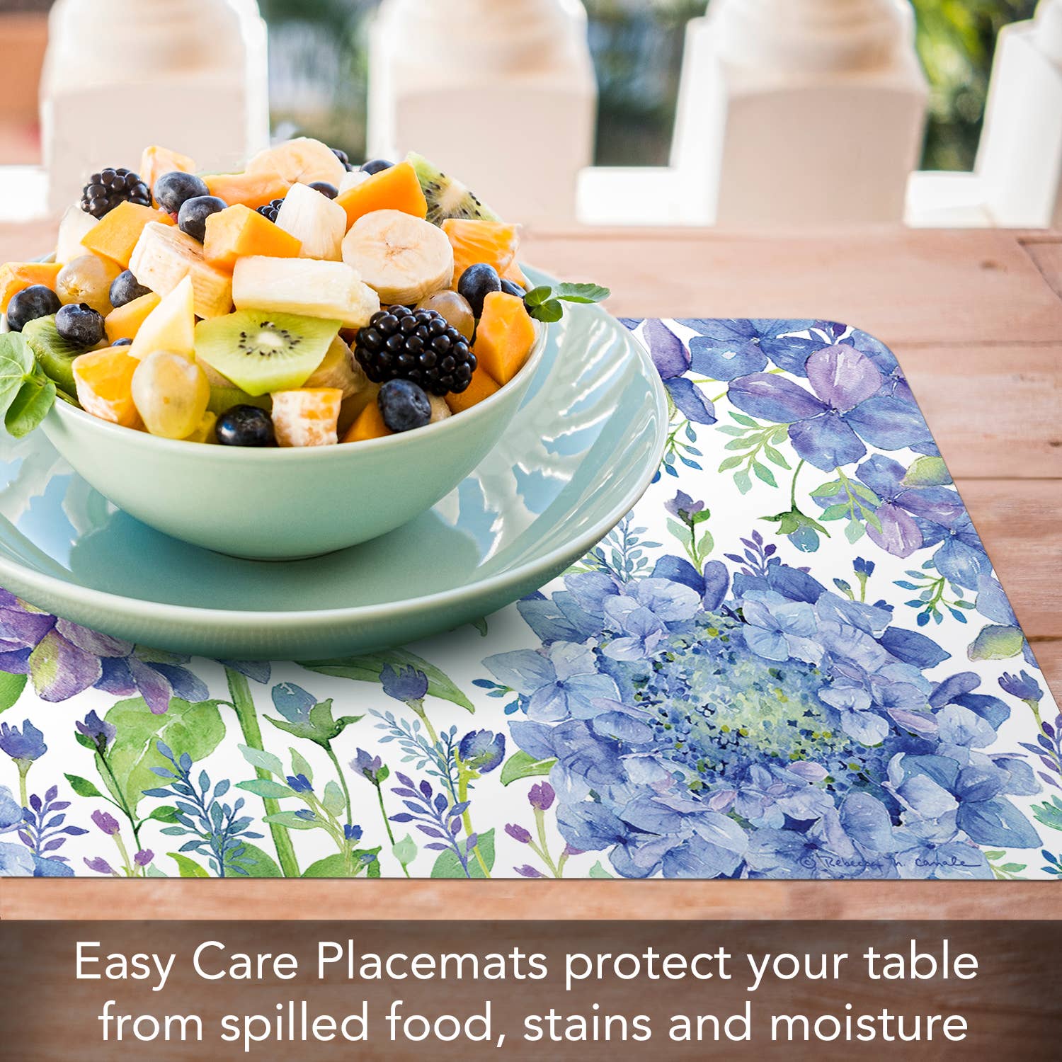 A bowl of mixed fruit salad on a blue plate rests on a floral placemat. Text below reads: Beautiful Hydrangea Reversible Rectangular Plastic Placemat, made from BPA-Free materials, protects your table from spills, stains, and moisture.