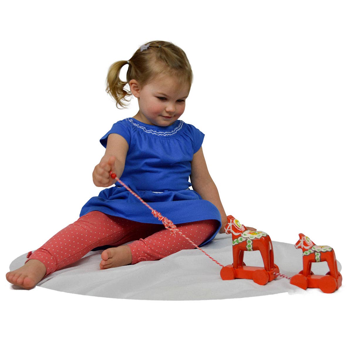 A young girl in a blue dress sits on a white mat, playing with the Swedish Horse Themed Pull Toy featuring two Dala horses, adding charming Swedish flair to her playtime.