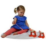 A young girl in a blue dress sits on a white mat, playing with the Swedish Horse Themed Pull Toy featuring two Dala horses, adding charming Swedish flair to her playtime.