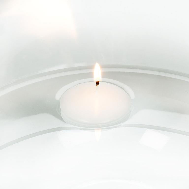 The 2" White Floater Tealight Candle is lit, casting a warm glow as it rests on a reflective glass surface, creating a cozy atmosphere.