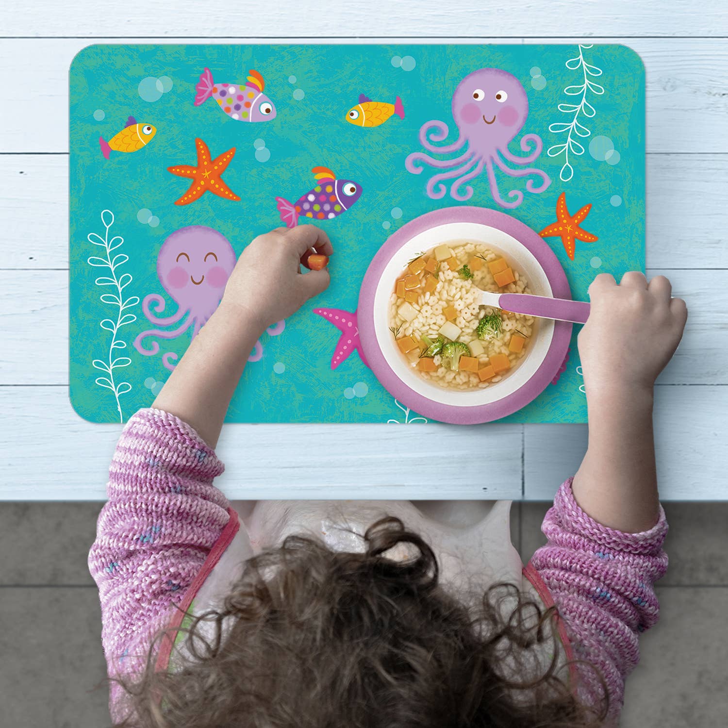 A child enjoys soup at a table adorned with the Kids Mermaid Reversible Rectangular Plastic Placemat, which features an ocean theme with fish, octopuses, and starfish. These BPA-free and easy-care placemats are a practical choice for parents and make an ideal housewarming gift.