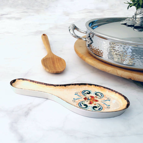 Ceramic spoon rest with a folk art floral pattern, placed beside a wooden spoon and a silver pot on a wooden trivet.