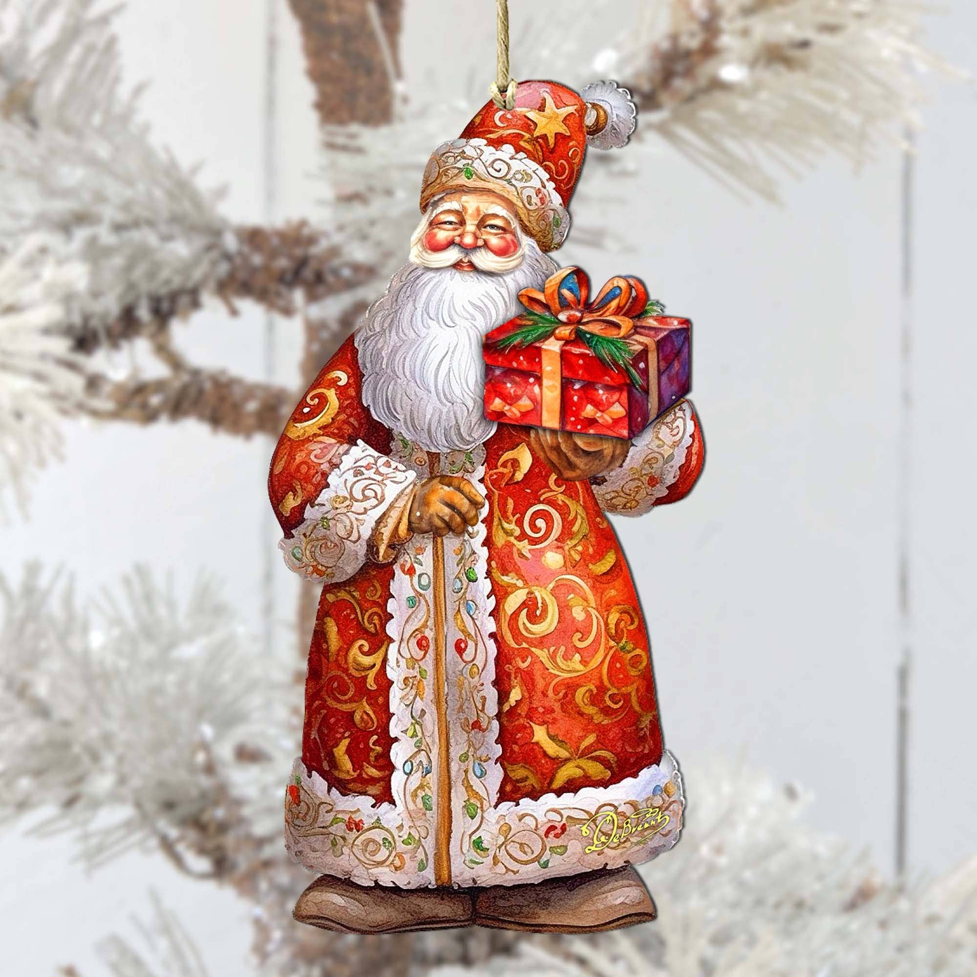 Introducing the St. Nick's Gift Wood Ornament from our Handcrafted Wooden Ornaments collection: this charming Santa Claus is adorned in a detailed red coat, holding a gift box as it gracefully hangs from a frosted Christmas tree branch.