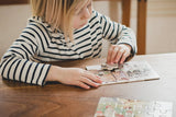 A child in a striped shirt sits at a wooden table, absorbed in assembling the Elsa Beskow "Children of the Forest" 4 Tray Puzzles, captivated by the delightful illustrations on each piece from the wooden box set.