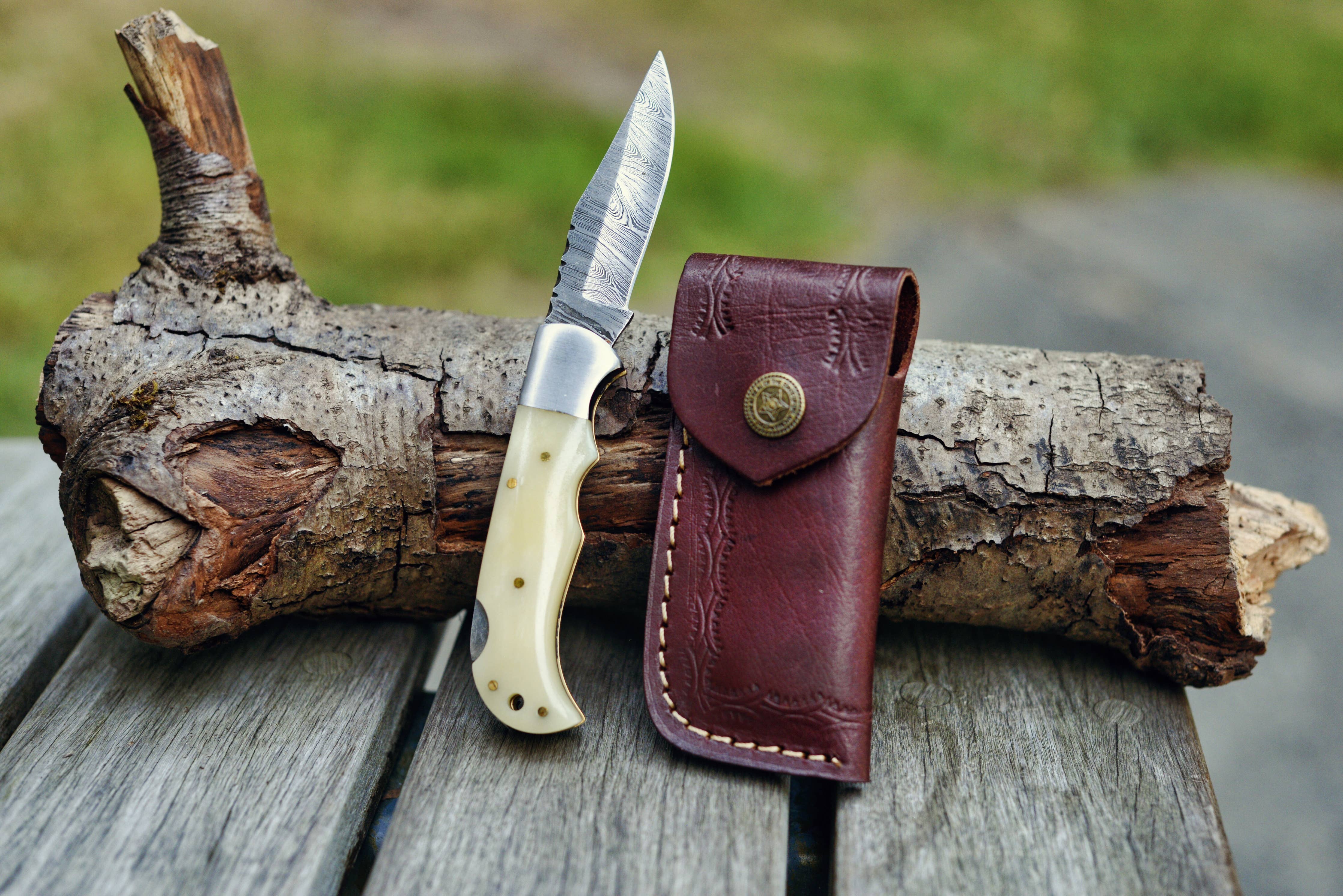 A Knife: Camel Bone Damascus Folding Knife, featuring a camel bone handle, is displayed against a piece of wood with its accompanying closed brown leather sheath on a wooden surface outdoors.