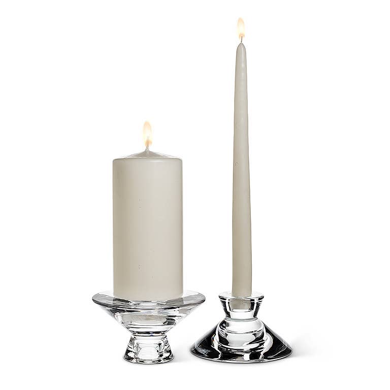 A pair of versatile Candle Holder: Reversible Glass Candleholders (4.5" D) elegantly showcases two lit white candles, one pillar and one taper. Their warm candlelight beautifully transforms any plain background into an inviting ambiance.