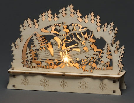 Laser cut wooden scene showing Santa in a sleigh pulled by reindeer, pine trees in the background, and a soft amber LED light illuminating the center.