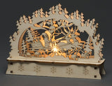 Laser cut wooden scene showing Santa in a sleigh pulled by reindeer, pine trees in the background, and a soft amber LED light illuminating the center.