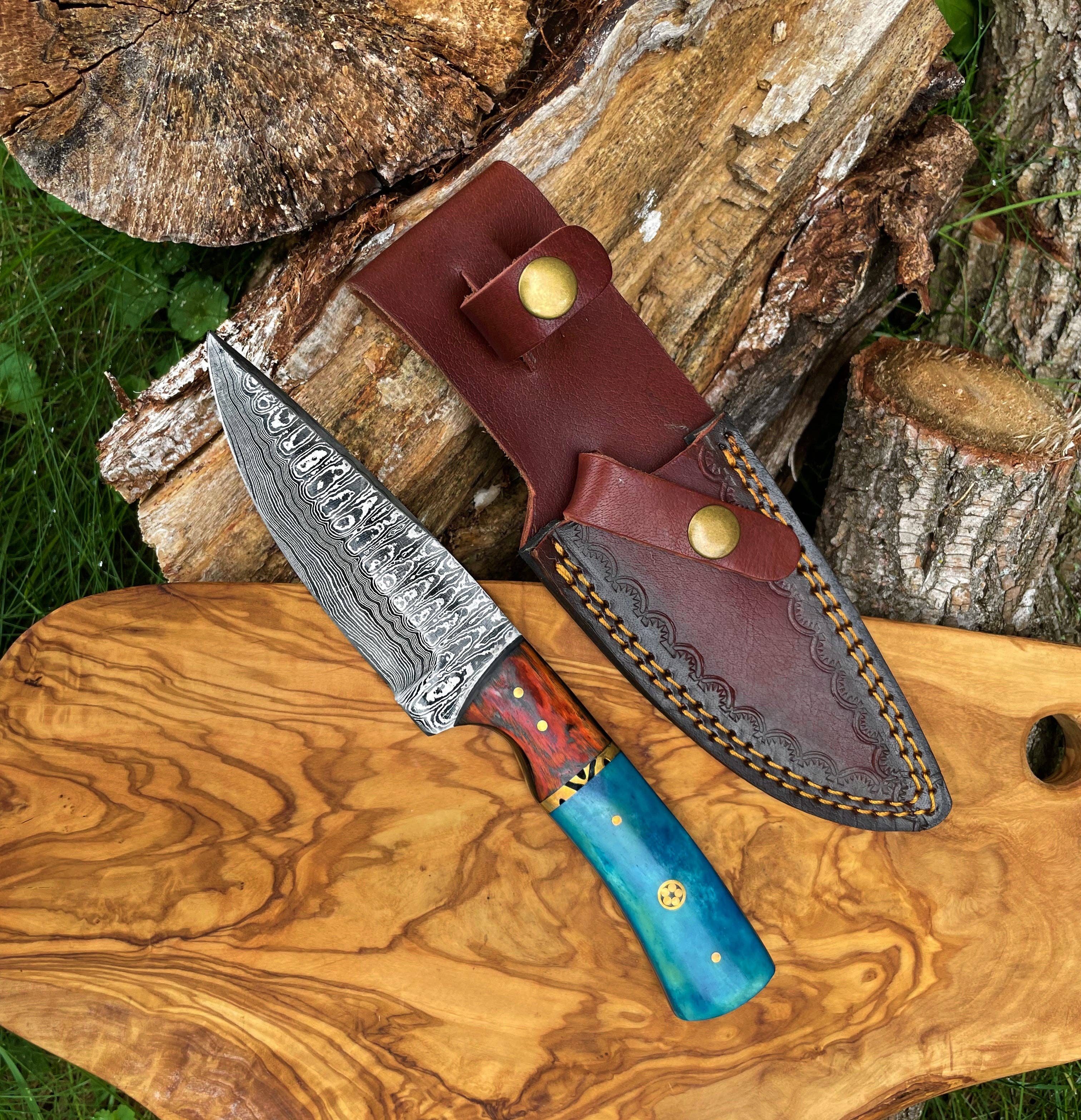 A Hand Forged Damascus Knife with a drop-style blade and black handle rests beside a brown leather sheath on a wooden surface, with logs in the background.
