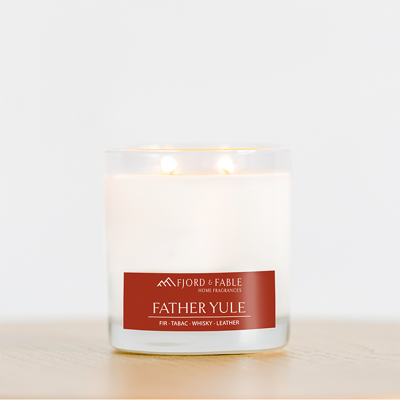 A white, two-wick soy candle in a 4oz jar by Fjord & Fable rests on a wooden surface. The label reads "Father Yule"—a festive scent of fir, tabac, whisky, and leather, with 20+ hour burn time for cozy evenings.