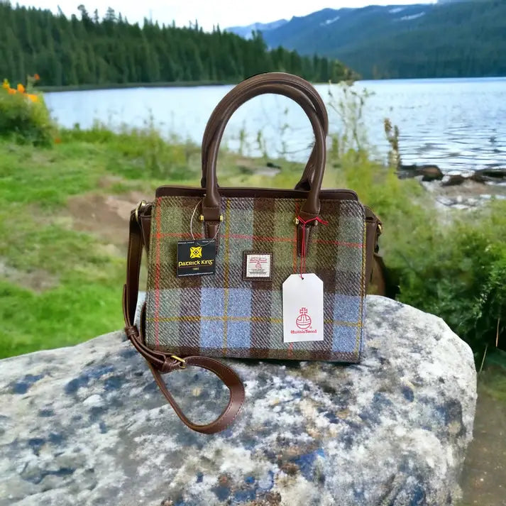 The Harris Tweed Double Handle Tote in dark brown, featuring plaid accents and tags, sits on a large rock overlooking a tranquil lake with trees and mountains in the background.