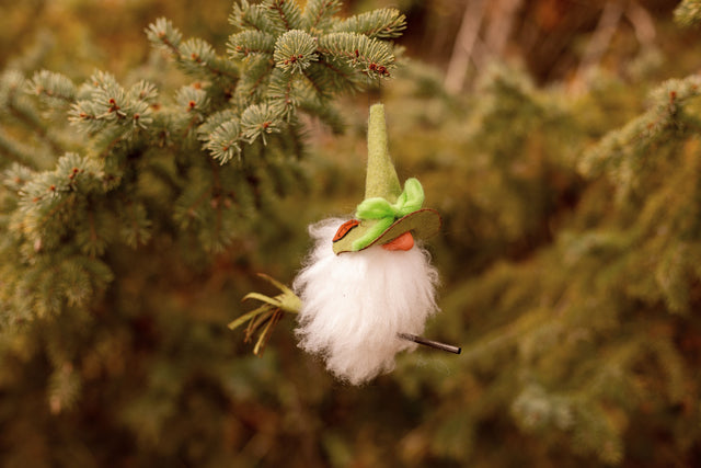 Scandinavian felt gnome ornament with a green hat and white beard, hanging from a pine branch.