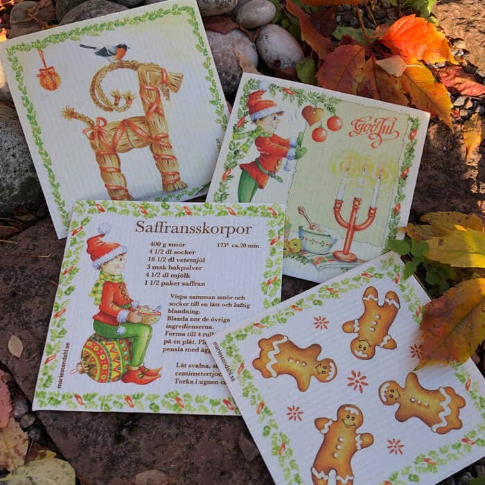 A Straw Goat with Red Ribbons Christmas dish cloth, featuring festive Swedish illustrations, is displayed among fallen autumn leaves and rocks.