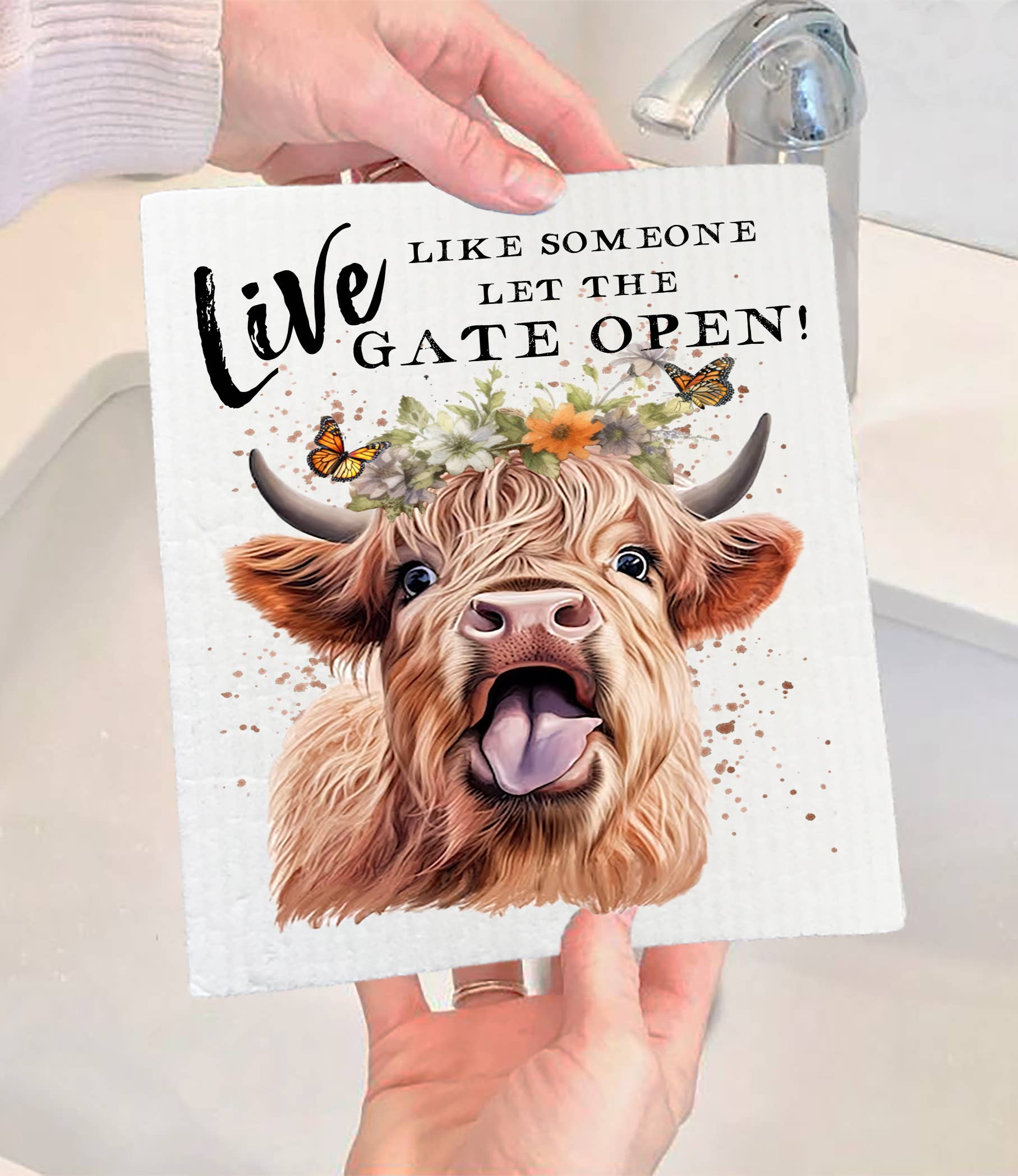 A person holds a Swedish Dish Cloth: Highland Cow Life Life Gate Open, featuring a cow with a flower crown and the text Live like someone let the gate open! This charming eco-friendly and compostable product is ideal for sustainable living.