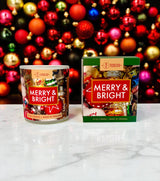 A Merry & Bright Candle 12oz and its box are displayed on a marble surface with festive ornaments, highlighting this premium holiday candle—ideal for adding a cheerful touch to your seasonal decor.