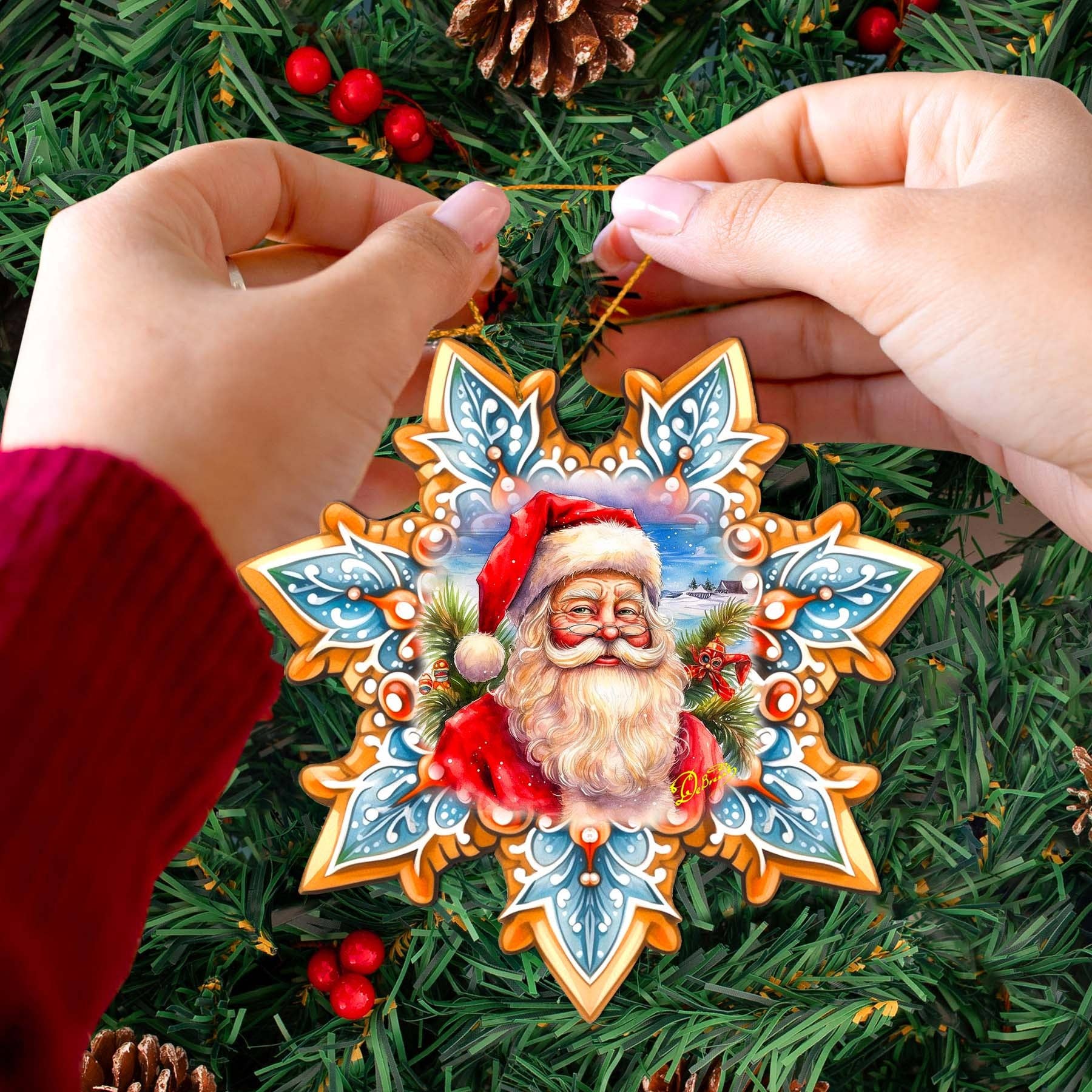 A person adorns their Christmas tree with a handcrafted Ornament: Santa Snowflake Wood Ornaments from GDebrekht, showcasing a charming depiction of Santa Claus amidst red berries and pinecones for that perfect holiday touch.