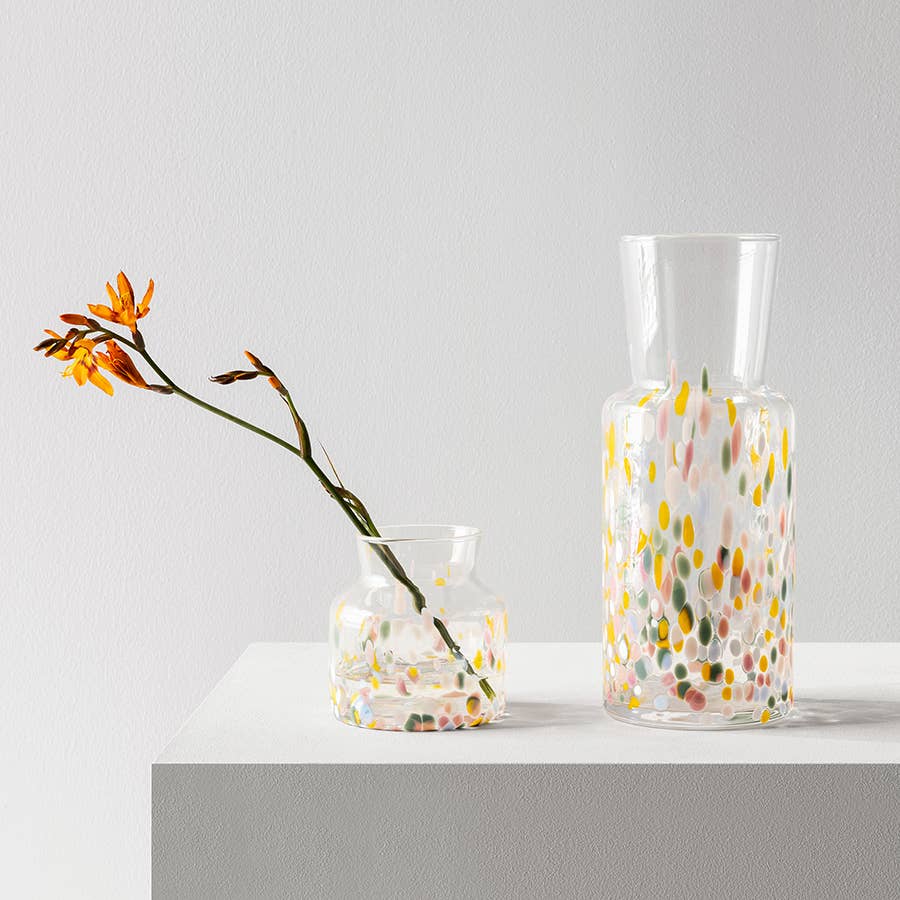 Two clear glass Meadow vases with multicolored confetti spots, one containing an orange flower.