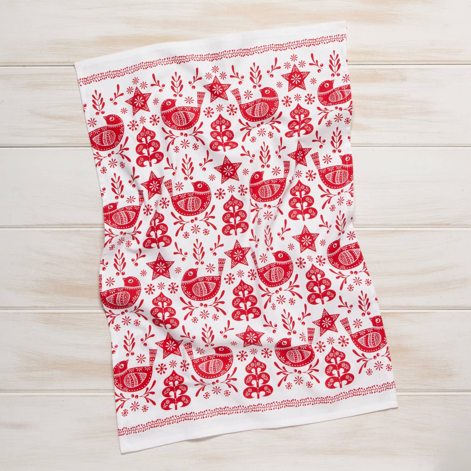 The Tea Towel: Snowbird Dishtowel, made entirely from 100% cotton, features a charming red and white design with stylized birds, trees, stars, and snowflakes. It's ideal for bringing a touch of elegance to your kitchen decor as it rests gracefully on a wooden surface.