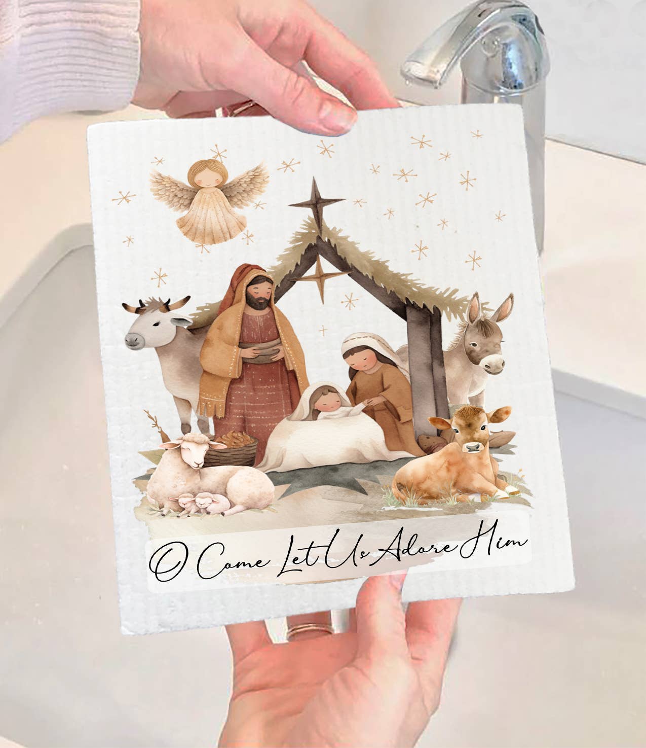 A person holds the "Dish Cloth: Christmas Nativity Christ is Born Swedish Dishcloth," which features Mary, Joseph, baby Jesus in a manger, animals, an angel, stars, and the text "O Come Let Us Adore Him.