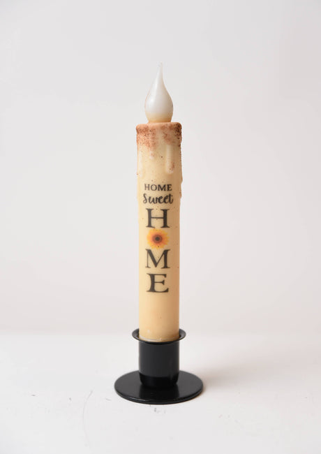 A tall cream-colored "Home Sweet Home Sunflower" LED timer taper candle, top dusted and battery operated, stands upright in a black holder against a plain white background.