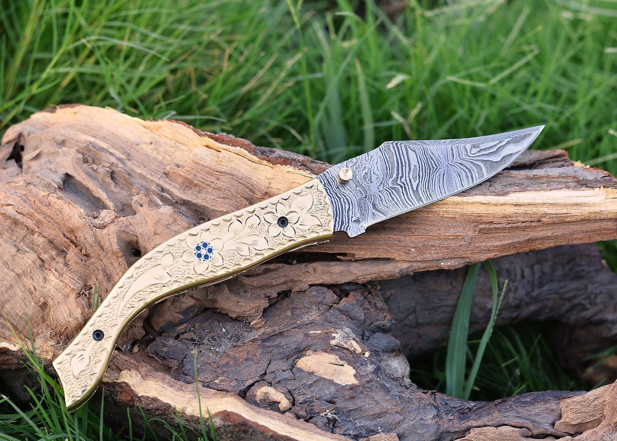 An Exquisite Golden Folding Knife with an engraved handle and stainless steel blade elegantly rests on wood atop the grass.