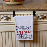 The "UFF Da" Norwegian-Swedish themed decorative tea towel hangs on a cabinet, featuring playful cartoons and text explaining "Uff Da"—a delightful Scandinavian kitchen gift.