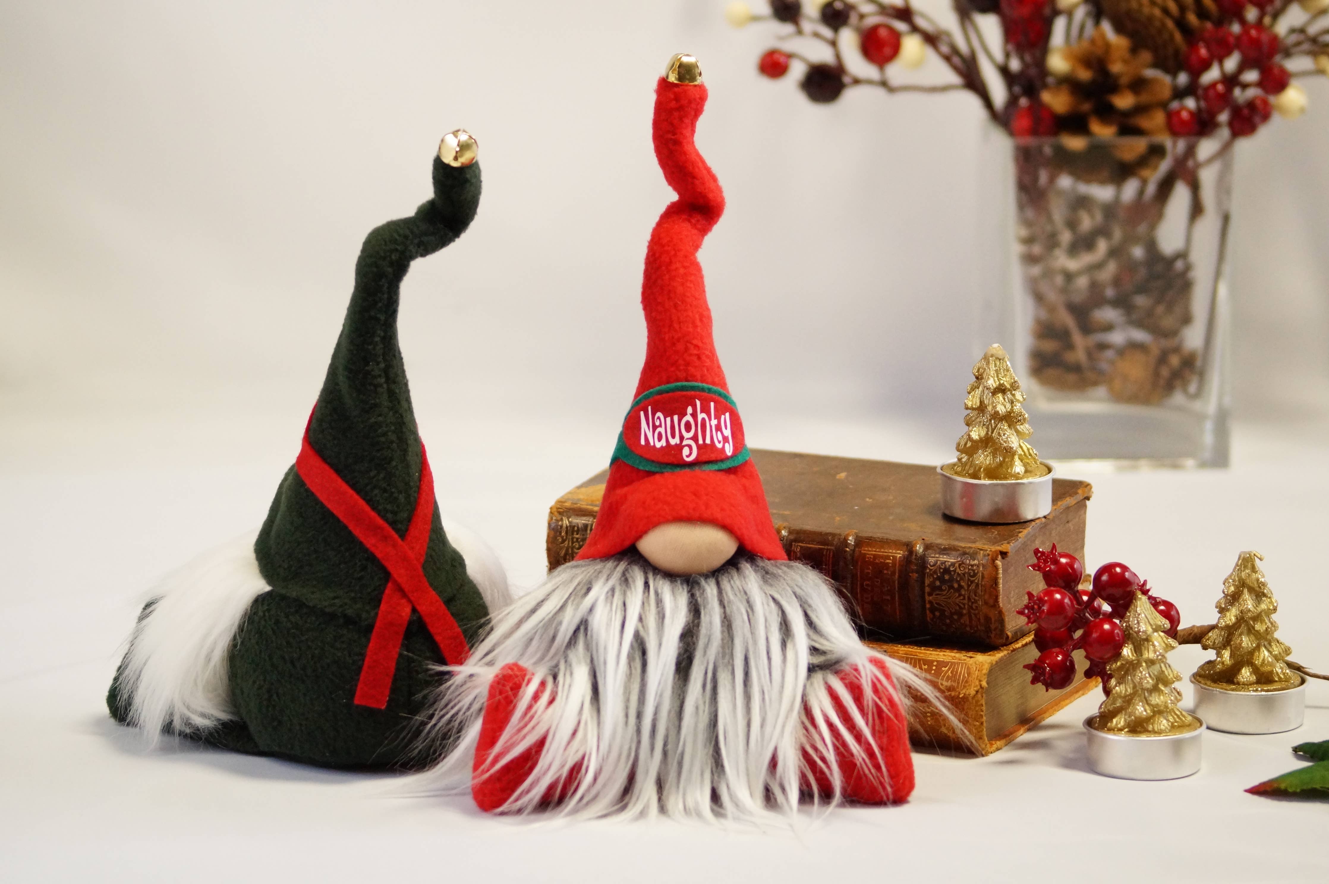 A pair of gnome figurines with red hats and long beards, featuring one branded as the Naughty Christmas Gnome, is displayed next to a stack of books, a small gold tree, and a vase filled with decorative berries.