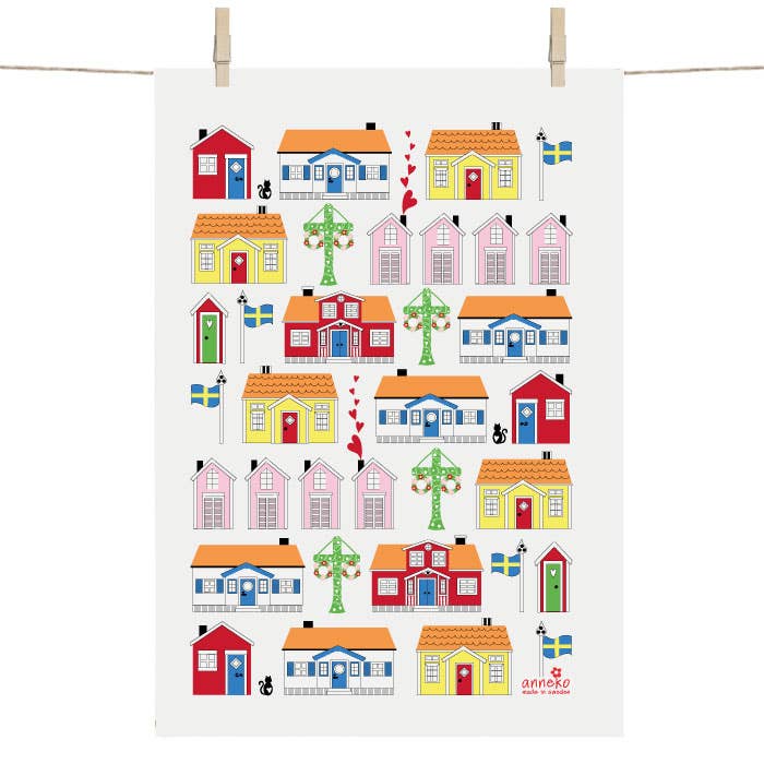 The Tea Towel - Swedish Cottages by Anneko of Sweden showcases colorful cottages, trees, flags, red hearts, and two black cats. Its beautifully designed and printed on a vertical sheet for hanging with clips.