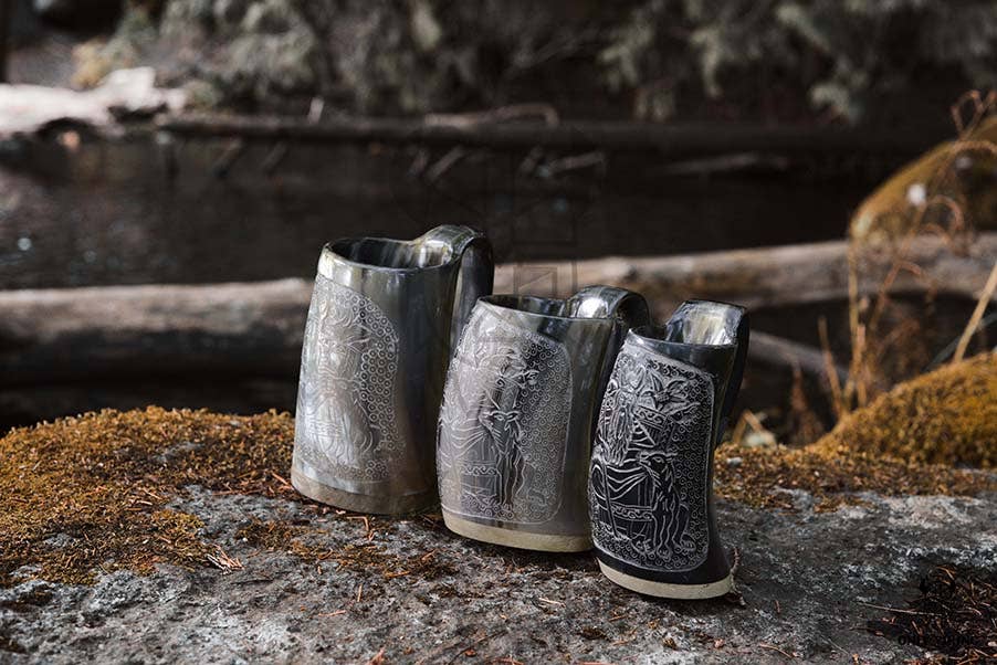 Three Horn: Viking Tankard pieces, each in white with black accents and intricately engraved, stand on a mossy rock by a calm stream, surrounded by blurred forest scenery.