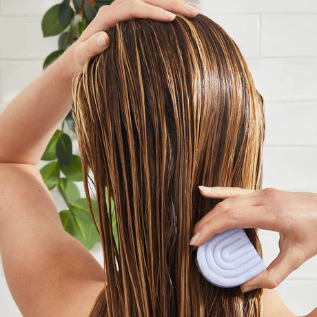 A person uses the Kitsch Purple Toning Shampoo & Conditioner Bar Combo for color-treated hair; a plant hangs in the background.