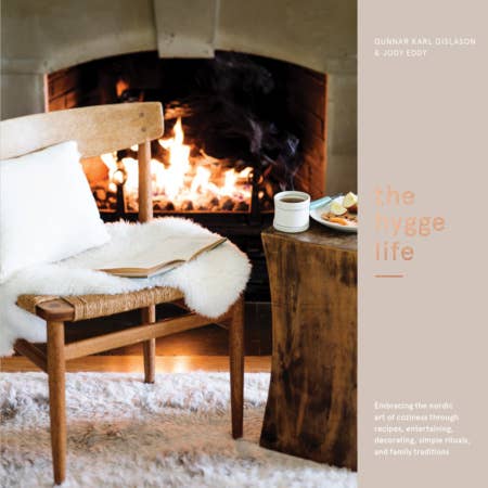 A wooden chair with a white cushion and sheepskin sits by the fire; a rustic table holds coffee, snacks, and the book "The Hygge Life" by Karl Gislason & Jody Eddy. Cozy recipes set the mood. Text reads: the hygge life.